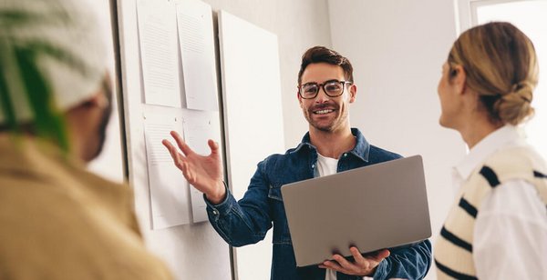 Mann mitt Laptop in der hand steht vor einem Memoboard und erklärt etwas